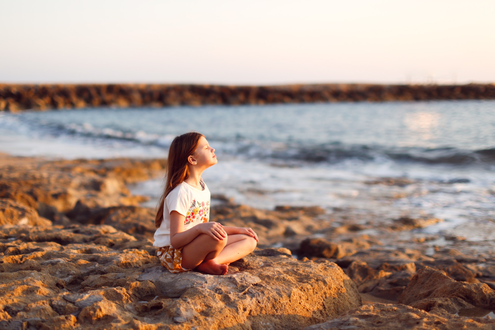 Girl kid meditates on a rock by the sea at sunset